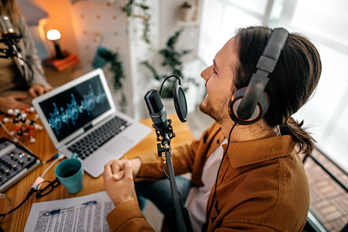 Handsome young men recording a podcast with notes using microphone, headphones and laptop in recording studio