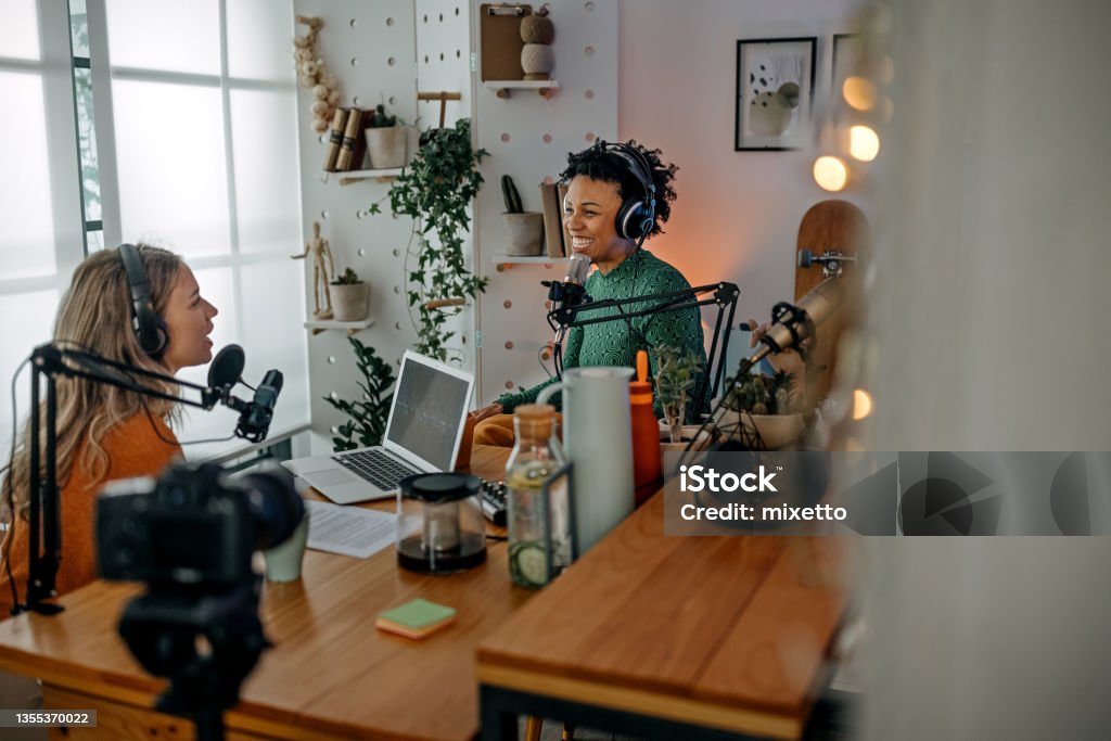 Two young women recording podcast Smiling young women recording podcast and interview with female guest using microphone and headphone in recording studio Podcasting Stock Photo