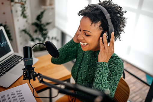 Smiling and successful young woman recording podcast on interview in studio using equipments like microphone, laptop and headphones