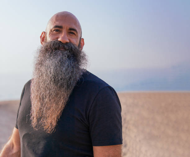Close-up portrait of a bald headed man with a long beard smiling and looking at the camera in a unfocused beach in the background.