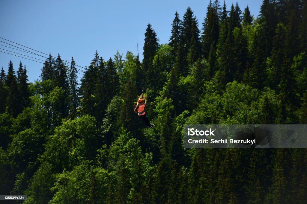 A girl of unrecognizable appearance descends a tightrope on a zipline at high altitude A girl of unrecognizable appearance descends a tightrope on a zipline at a high altitude. Attraction on the background of mountains covered with forest Zip Line Stock Photo