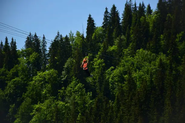 A girl of unrecognizable appearance descends a tightrope on a zipline at a high altitude. Attraction on the background of mountains covered with forest