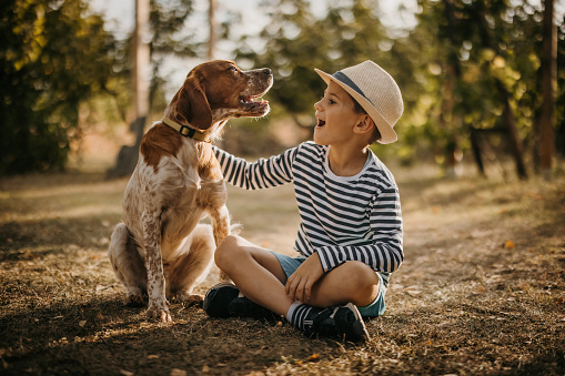Cute little boy with hat sitting on grass in back yard and stroking his dog