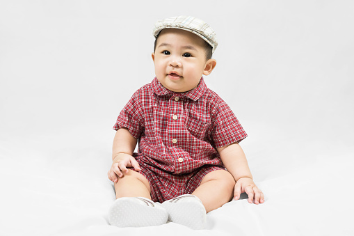 Cute 6-7 months little asian baby boy in casual outfit and hat sitting on white blanket cool and happily at home,Stylish fashionable kid. Adorable child in fashion and Style