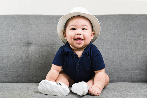 Adorable baby boy  first sitting on sofa. Cute infant Asian about 5-6 months old wearing blue t-shirt,white hat and shoes smiling and looking at camera.