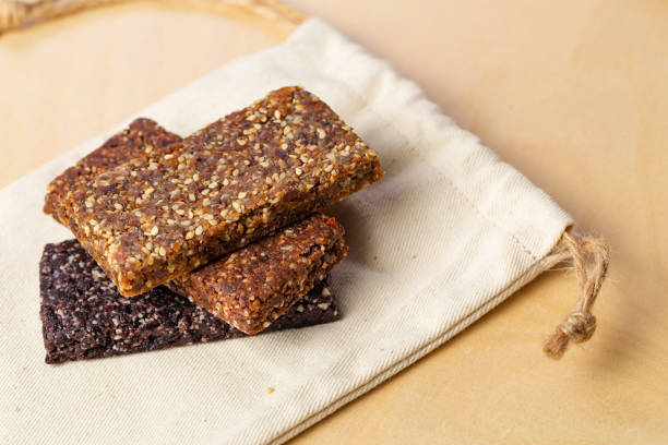 Homemade organic granola bars made by hand with fine grinded nuts and dry fruits piled on top above each other. Macro image with selective focus, view from above on wooden creamy surface and light textile bag.
