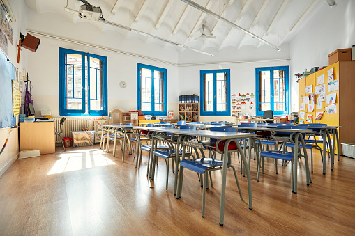 Unoccupied learning environment with hardwood floor, windows providing natural light, chalkboard, and child-size desks and chairs.