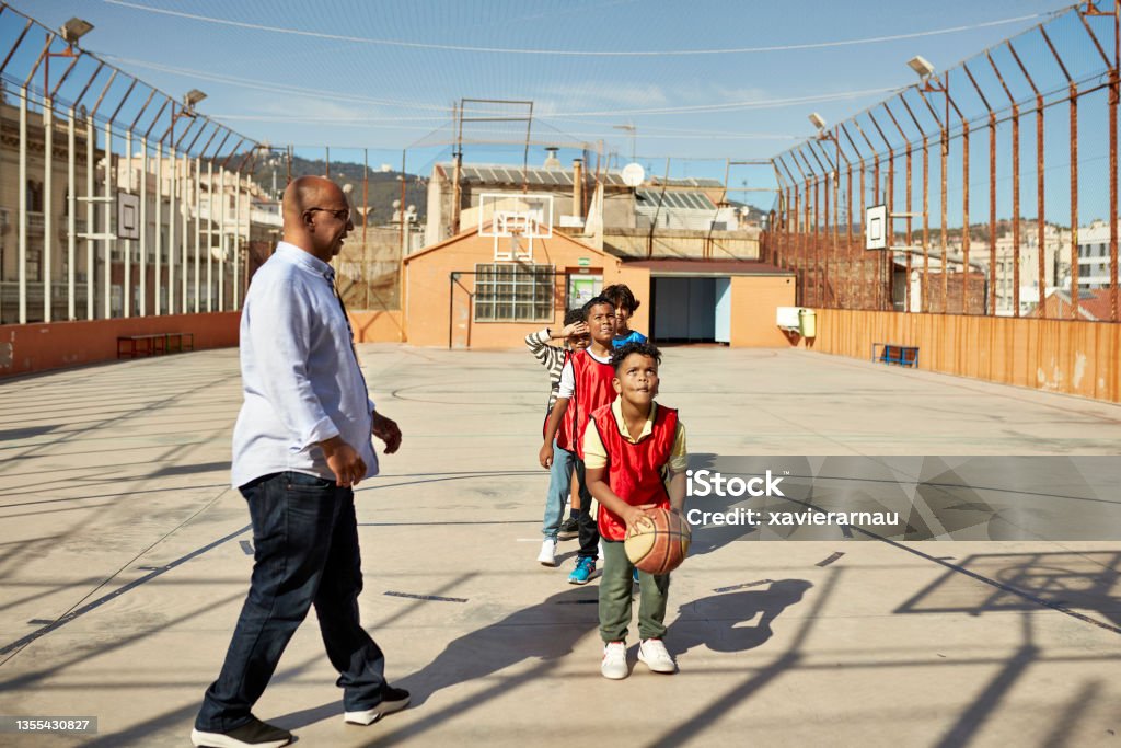 Young Schoolboy Concentrating Before Shooting a Free Throw Elementary age boys in red scrimmage vests waiting in line for a chance to take a basketball shot under the guidance of male teacher in late 40s. Basketball - Sport Stock Photo
