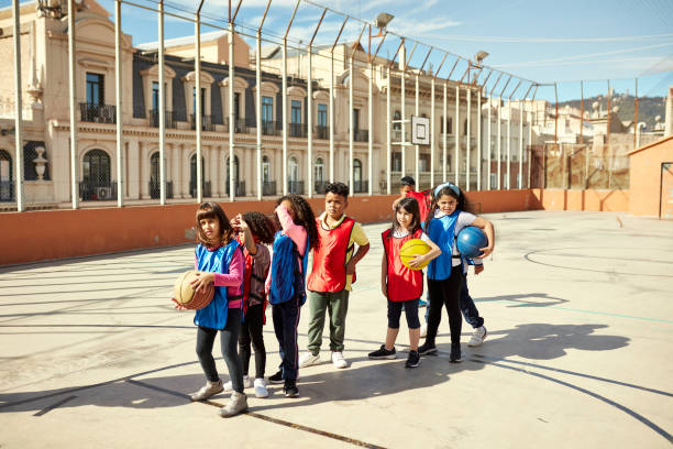 Schoolgirls and boys in blue and red scrimmage vests holding basketballs for a turn to take a shot at the basket.