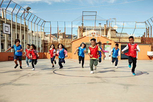 Wide angle front view of 10 energetic schoolchildren in red and blue scrimmage vests racing each other from one end of the court to the other during playtime.