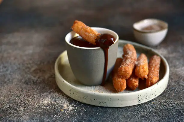 Traditional spanish dessert churros - fried choux pastry with chocolate sauce on a plate on a dark slate, stone or concrete background.