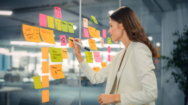 istock Young Confident Businesswoman Creating Project Plan on Office Wall with Paper Notes. Stylish Beautiful Manager Working on Business, Financial and Marketing Projects. Specialist in Diverse Team. 1355440522