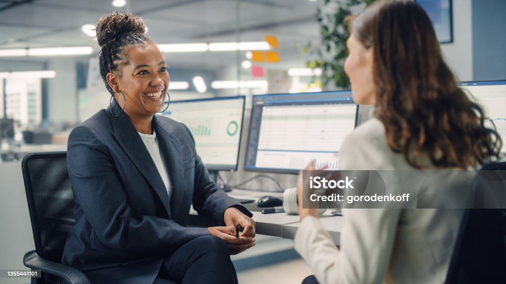 Two Female Colleagues Fondly Talk to Each Other, Laugh and Smile while Working on Computers in Diverse Modern Business Office. Experienced Manager and Young Employee Discuss a Fun Analytical Project. Manager Stock Photo
