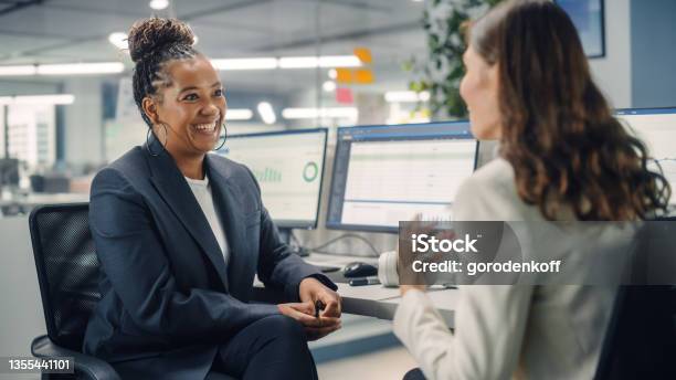 Two Female Colleagues Fondly Talk To Each Other Laugh And Smile While Working On Computers In Diverse Modern Business Office Experienced Manager And Young Employee Discuss A Fun Analytical Project Stock Photo - Download Image Now