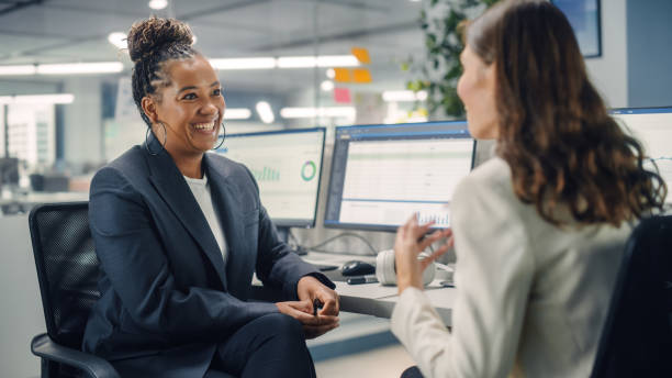 two female colleagues fondly talk to each other, laugh and smile while working on computers in diverse modern business office. experienced manager and young employee discuss a fun analytical project. - spoke imagens e fotografias de stock