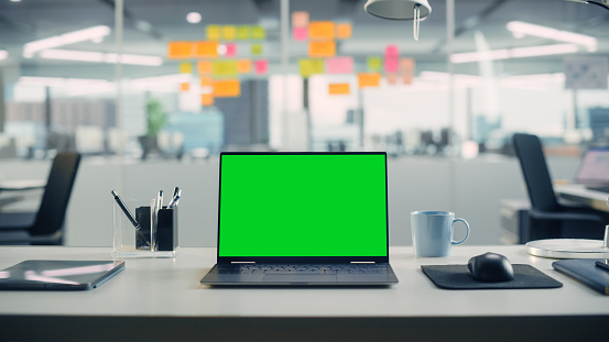 Laptop Computer with Mock Up Green Screen Chroma Key Display Standing on the Desk in the Modern Creative Office. In the Background Glass Wall with Colorful Planning Notes.