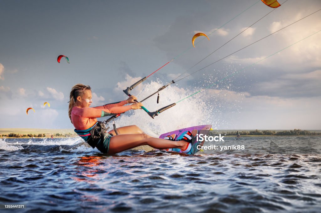 Professional kitesurfer young caucasian woman glides on a board along the sea surface at sunset against the backdrop of beautiful clouds and other kites. Active water sports Professional kitesurfer young caucasian woman glides on a board along the sea surface at sunset against the backdrop of beautiful clouds and other kites. Active water sports. Kiteboarding Stock Photo