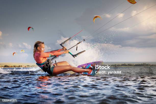 Professional Kitesurfer Young Caucasian Woman Glides On A Board Along The Sea Surface At Sunset Against The Backdrop Of Beautiful Clouds And Other Kites Active Water Sports Stock Photo - Download Image Now