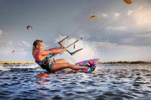 Professional kitesurfer young caucasian woman glides on a board along the sea surface at sunset against the backdrop of beautiful clouds and other kites. Active water sports.