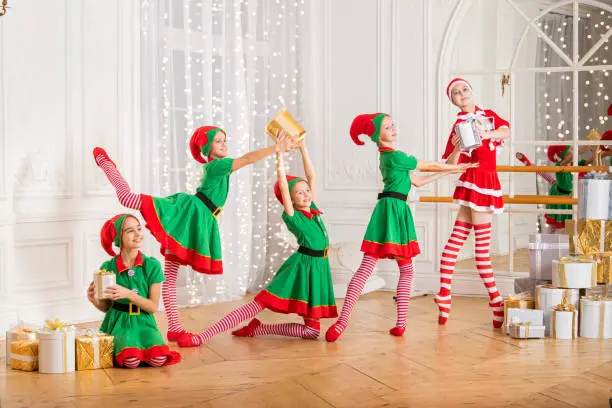 Photo of Children dressed as elves and practicing at a ballet barn look at a dancing ballerina dressed as santa claus in a spacious white studio.