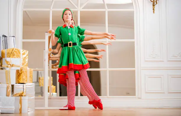Photo of Children dressed in costumes of Christmas elves practice at a ballet bar in front of a mirror in a spacious white studio.