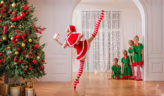 Children dressed as elves watch dancing santa claus ballerina near a Christmas tree in spacious white studio.