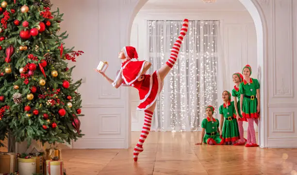 Photo of Children dressed as elves watch a dancing santa claus ballerina near a Christmas tree in a spacious white studio.