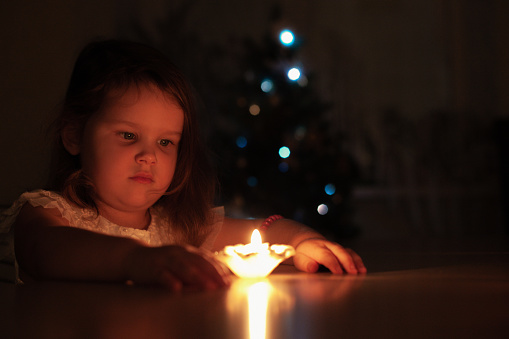 portrait of thoughtful, four-year-old girl looking at candle on table and making wish at home at Christmas night with Christmas tree on background.