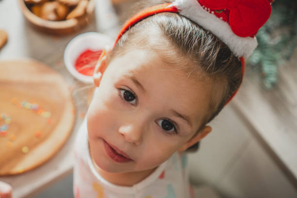 a garotinha de cabelos escuros de 3 anos de idade na faixa de cabelo do papai noel olha para a câmera na cozinha. feliz natal. feliz ano novo - preschool 2 3 years preschooler little girls - fotografias e filmes do acervo