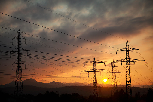Pylons of a high voltage power line at dawn, wires and poles. Orange sun over the land. Evening sun.