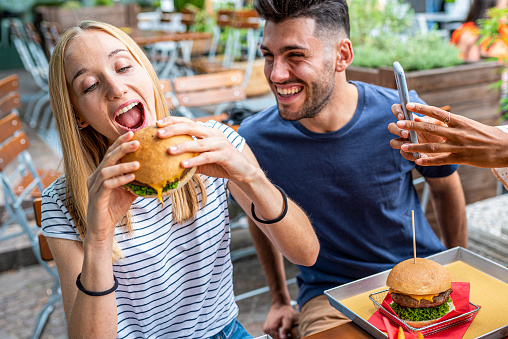 Group of friends having fun while eating at fast food, happy people smiling and laughing at hamburgers restaurant, millennials eating junk food