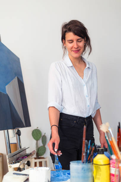 istock In her studio, a young smiling painter smears paint with a paintbrush on a palette. Around it is a work of art placed on an easel, as well as other accompanying material for painting. 1355459493