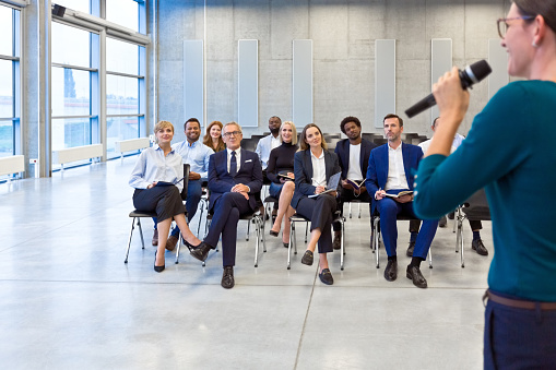 Multiracial group of business people sitting on chairs in the convention center during business meeting, listening to female speaker.