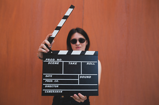 A young girl with sunglasses and piercings holding a movie clapboard in front of a orange background. Movie film director concept.