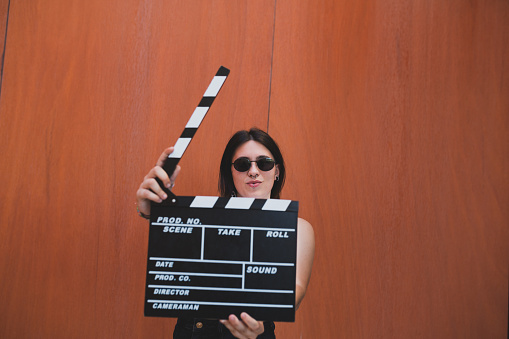 A young girl with sunglasses and piercings holding a movie clapboard in front of a orange background. Movie film director concept.