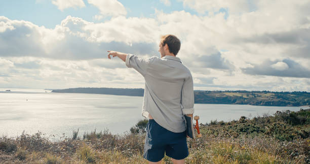 istock Young man with skateboard under arm walking at beautiful ocean cost pointing at something 1355473537