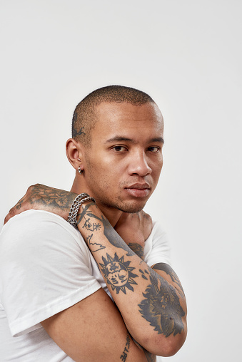 Portrait of serious young mixed race tattooed guy in white t shirt posing for camera isolated over white background. Freestyle, human emotions, youth concept