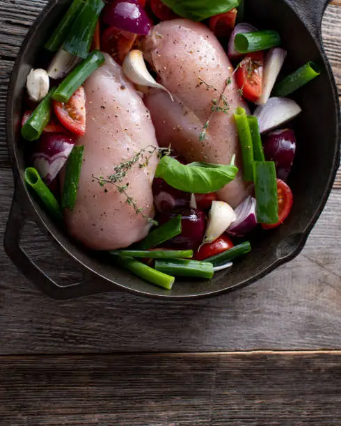 Uncooked chicken breast with raw vegetables in a rustic pan isolated on wooden background. Overhead view with copy space