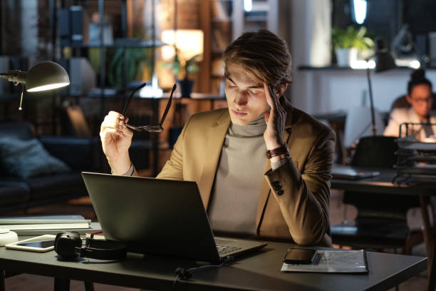istock Overworked young manager massaging temple while feeling headache after long work on laptop in dark office 1355489206