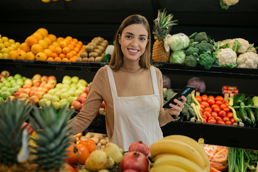 Portrait of a happy young Caucasian woman at a grocery store, working next to the fruits and vegetables, checking the quality of the items using a smart phone