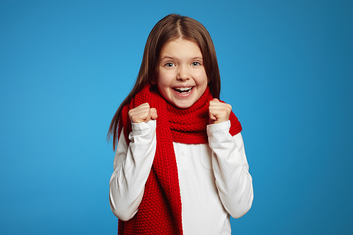 Cheerful little girl wearing red scarf, raising fists up and celebrating while looking at camera, isolated over blue background