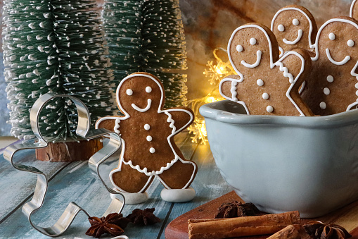 Stock photo showing close-up view of Christmas scene of homemade gingerbread men iced with white, royal icing sat 'tobogganing' in a ceramic cake pan, surrounded by model fir trees and illuminated fairy lights.
