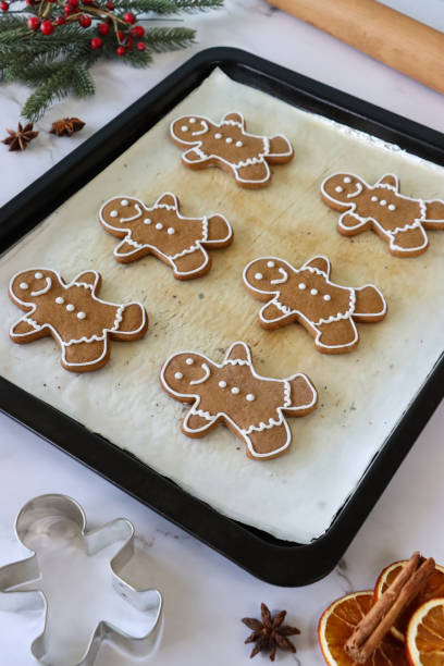 Stock photo showing close-up, elevated view of marble effect background with batch of homemade gingerbread men decorated with white, royal icing in baking tray, lined with greaseproof parchment paper. imagen de bandeja para hornear, forrada con papel de pergamino a prueba de grasa de casera, galletas de hombre de pan de jengibre heladas con blanco, glaseado real, rodillo, cortador de galletas, rodeado de palitos de canela, rodajas secas de cítricos ana