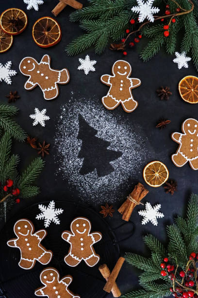 istock Image of Christmas tree stencilled, white icing sugar on black background, surrounded by gingerbread men cookies, fondant icing snowflakes, dried citrus orange fruit slices, star anise, cinnamon sticks, spruce needles and red berries, elevated view 1355500701