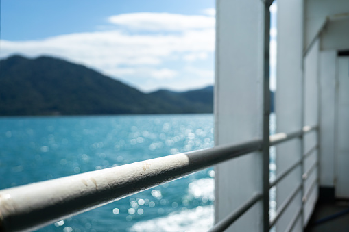 Close-up a white falling prevention rail at corridor walkway of the passenger boat with blurred background of ocean and island view. Transportation and travel scene. Selective focus at railing part.