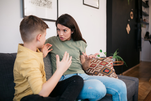 Young Caucasian teenage boy sitting on a sofa while his mother looks angry and is arguing with him