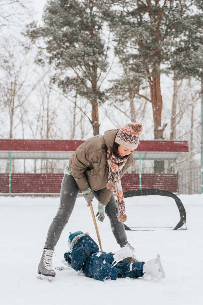 mały zabawny chłopiec jeżdżący na łyżwach w parku i upadł z matką. naucz się grać w hokeja na lodzie z kijem. odkryty. sporty zimowe - ice skating ice hockey child family zdjęcia i obrazy z banku zdjęć