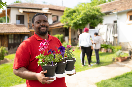 An Jamaican-British man is smiling at the camera while he is standing in his garden holding three plant pots full of flowers, his family are in the garden behind him