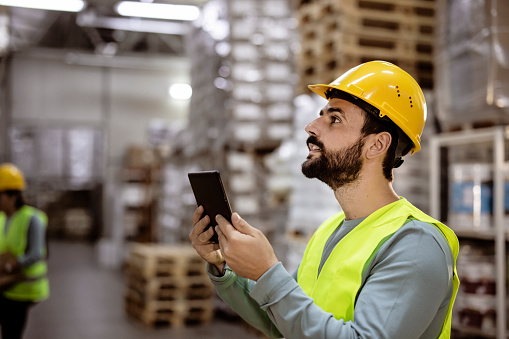 Young Male Factory Manager Using Digital Tablet in Warehouse While Standing Against Goods Shelf