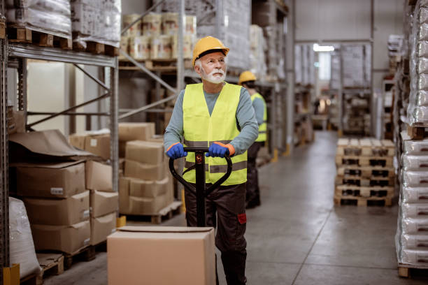Keeping up with the movement of stocks Senior male worker in uniform in the warehouse with pallet truck cardboard helmet stock pictures, royalty-free photos & images